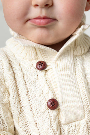 Close-up of a child wearing a cream-colored cable knit sweater with a mock neck and brown buttons. The sweater is made from soft, sustainable organic cotton, perfect for young kids.