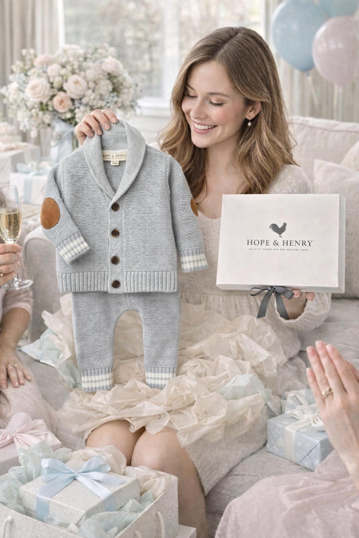 A woman in a light sweater holds a gray heather baby cardigan and legging set from Hope & Henry, surrounded by friends and gift boxes in a cozy, festive setting.
