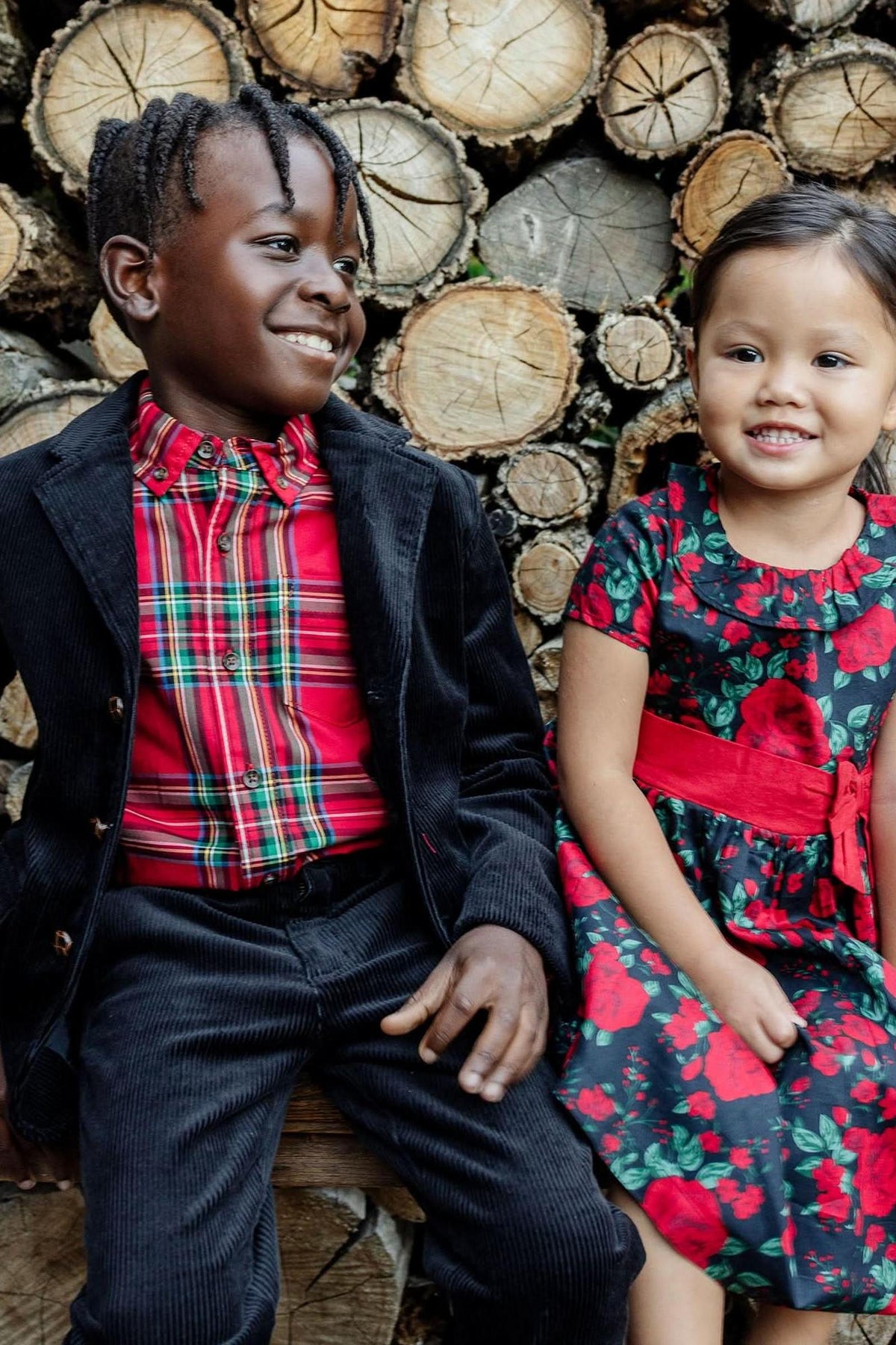 A boy in a plaid shirt and corduroy suit sits beside a girl in a floral dress with a red.