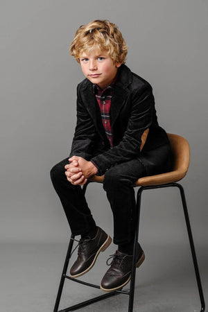 A young boy sits on a stool, wearing black corduroy pants and a plaid shirt, with stylish brown shoes.