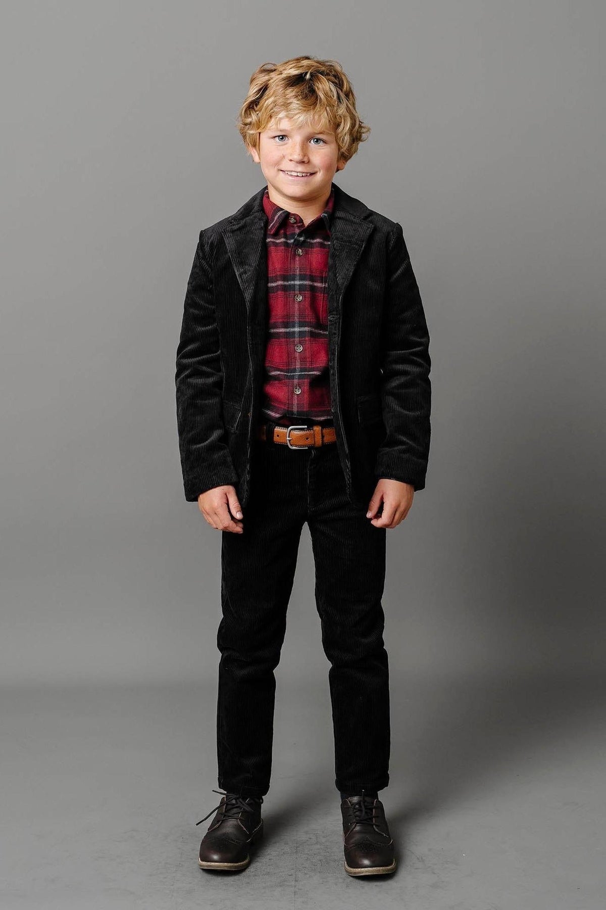 A smiling boy wears a black corduroy suit jacket over a red plaid shirt, paired with black corduroy pants and.