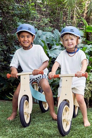 Two children are smiling while riding wooden balance bikes in a lush green garden. Each child wears a blue helmet, showcasing a fun and safe outdoor play environment.