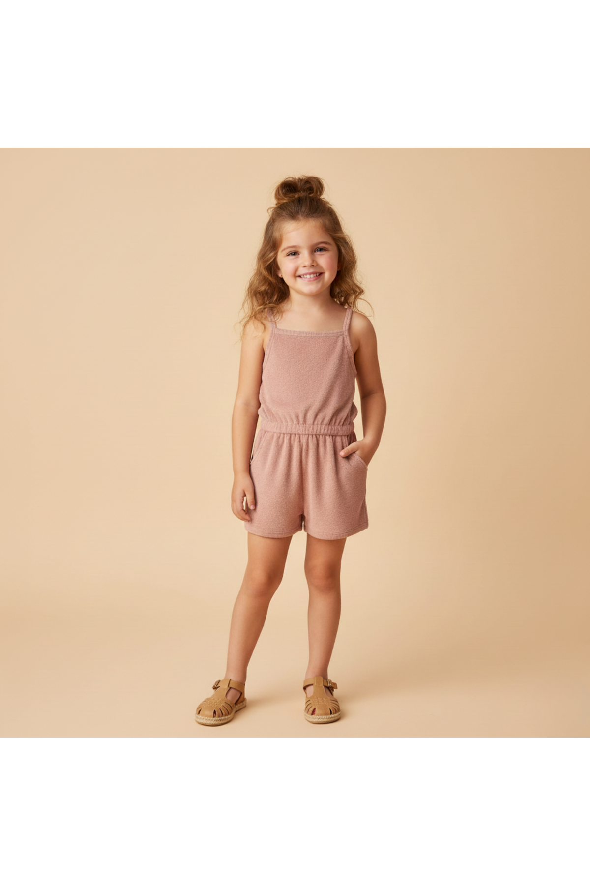 A smiling young girl stands in a light clay-colored organic terry tank romper with an elastic waist and pockets, set against a soft beige background, perfect for warm-weather play.