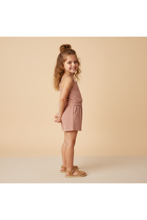 A young girl stands sideways, smiling while wearing a clay-colored organic tank romper made from soft terry fabric. The backdrop is a warm, neutral tone, enhancing the rompers casual style.