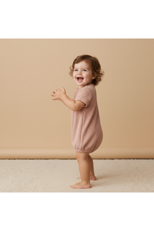 A joyful toddler with curly hair stands on a soft surface, wearing a clay-colored organic cotton romper. The relaxed fit and playful style are perfect for warm-weather adventures.