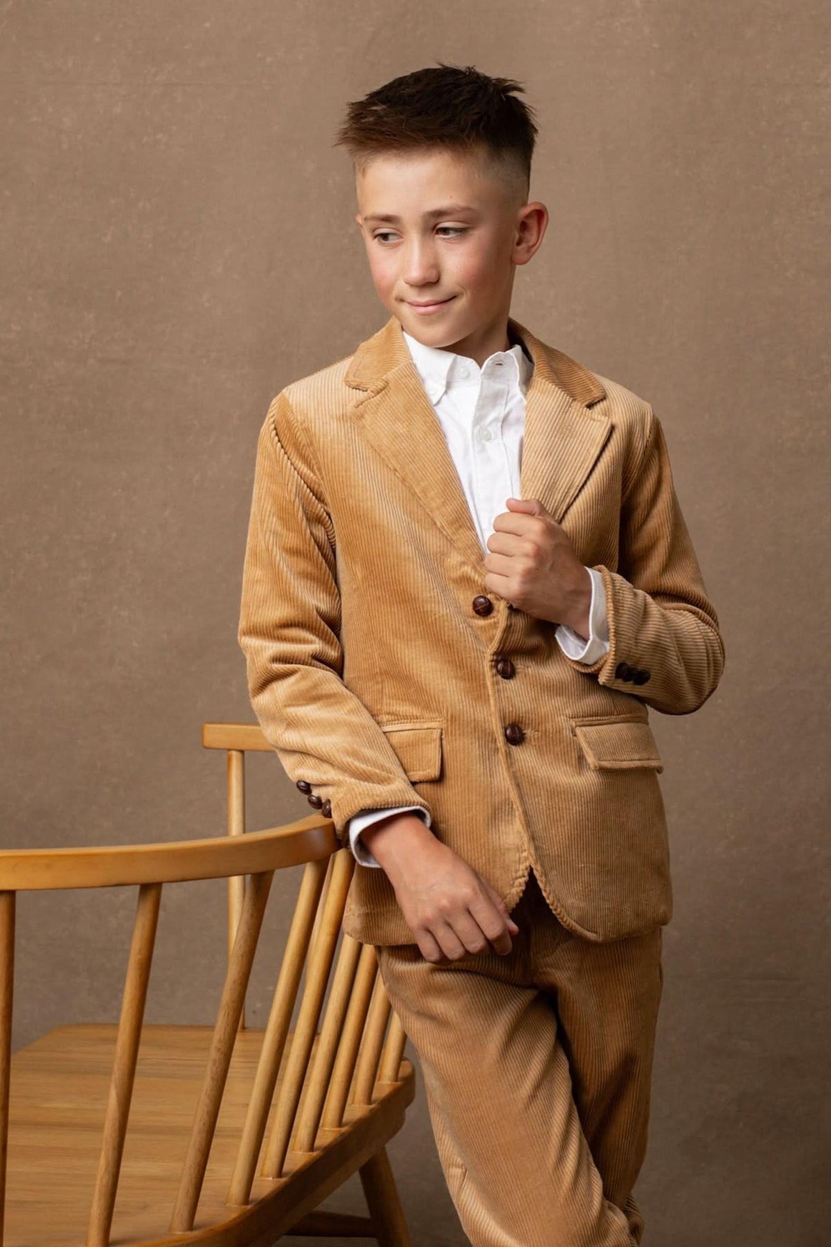 A young boy models a medium brown organic corduroy blazer, featuring button details and a tailored fit, paired with a white shirt.