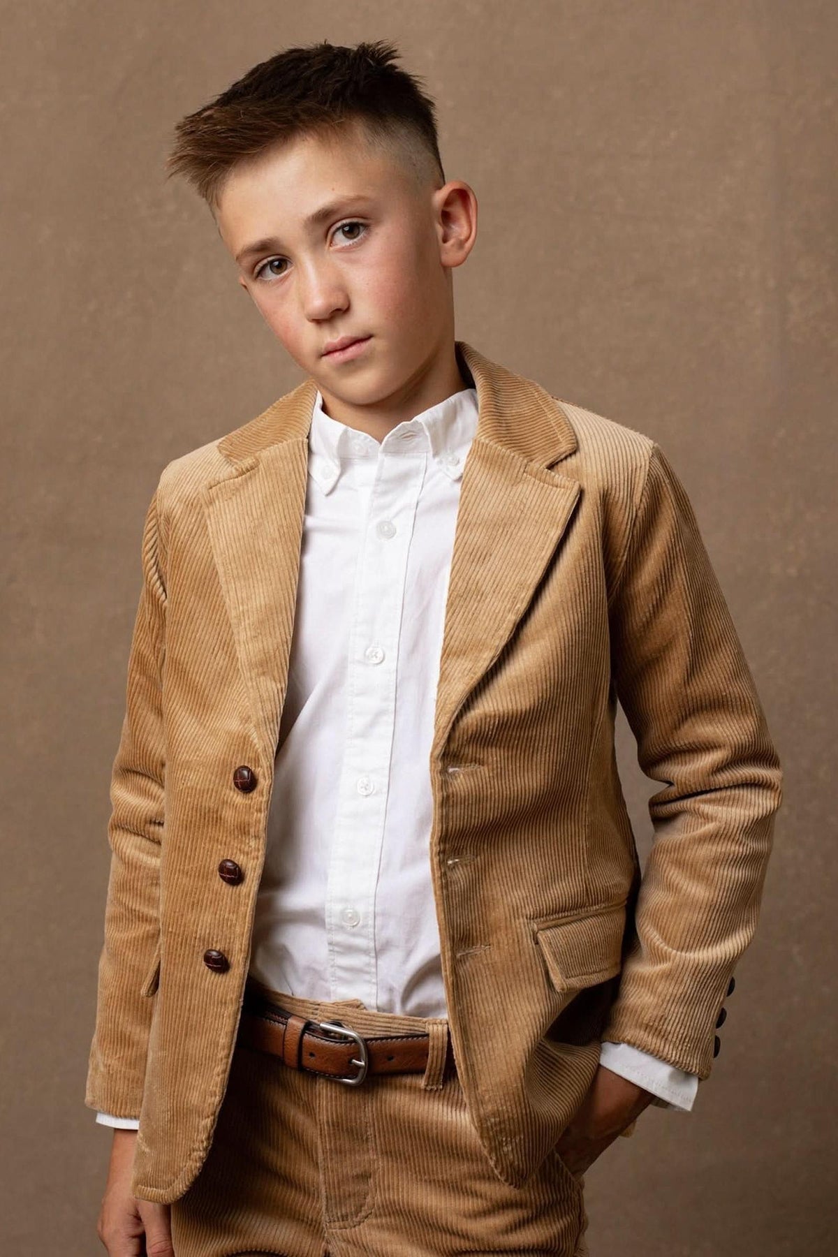 A young boy stands confidently in a medium brown corduroy blazer, featuring button details and a classic cut, paired with a white dress shirt against a neutral backdrop.