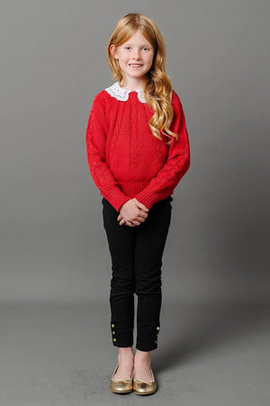 A young girl stands confidently in a red cable-knit sweater with a white collar, paired with black ponte pants featuring button cuffs. She completes her look with shiny gold flats.