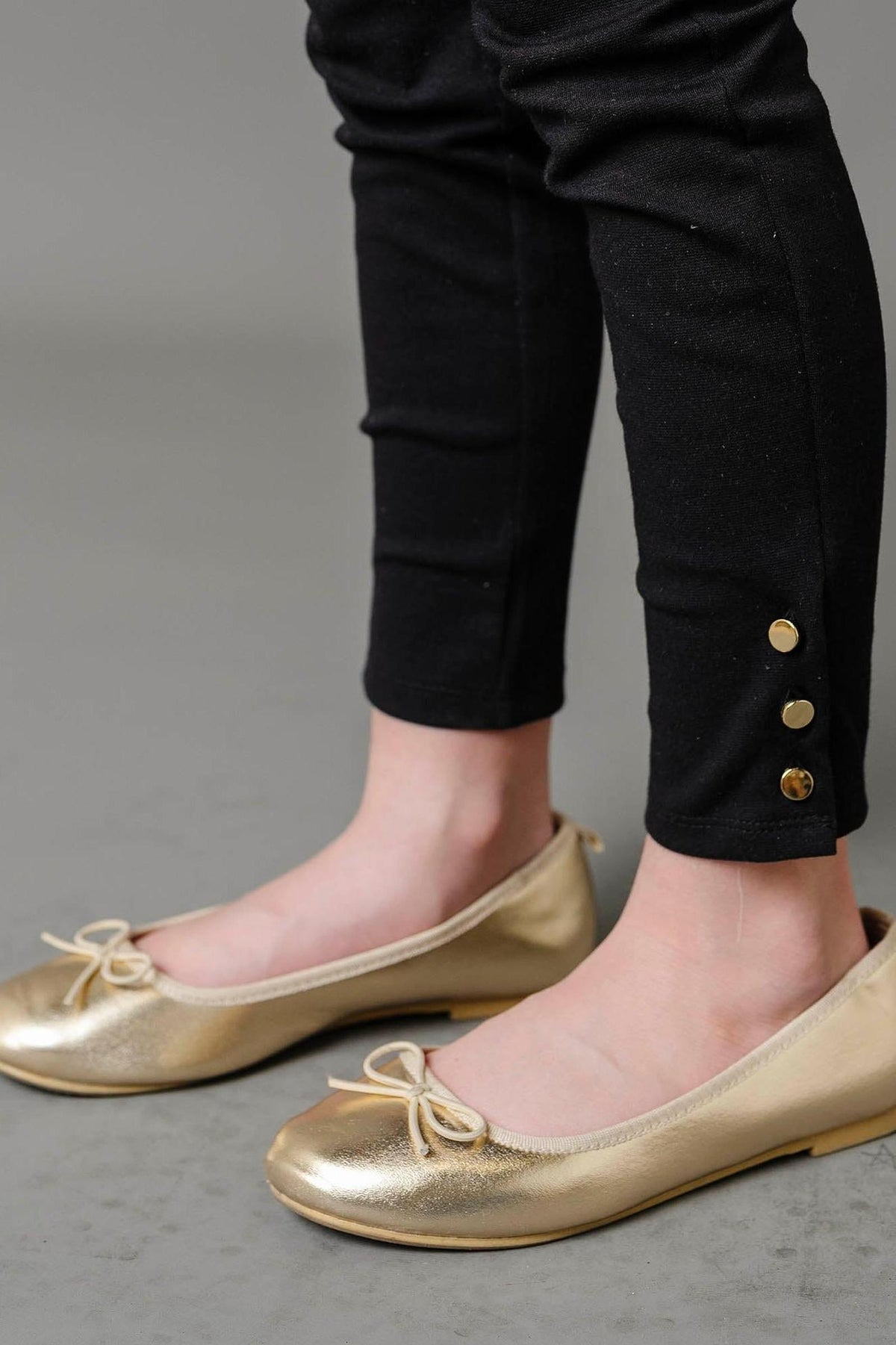 A close-up of a childs legs wearing black ponte pants with gold button cuffs, paired with shiny gold ballet flats. The background is a neutral gray, emphasizing the stylish outfit.