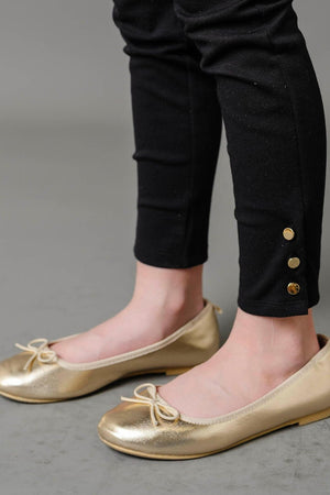 A close-up of a childs legs wearing black ponte pants with gold button cuffs, paired with shiny gold ballet flats. The background is a neutral gray, emphasizing the stylish outfit.