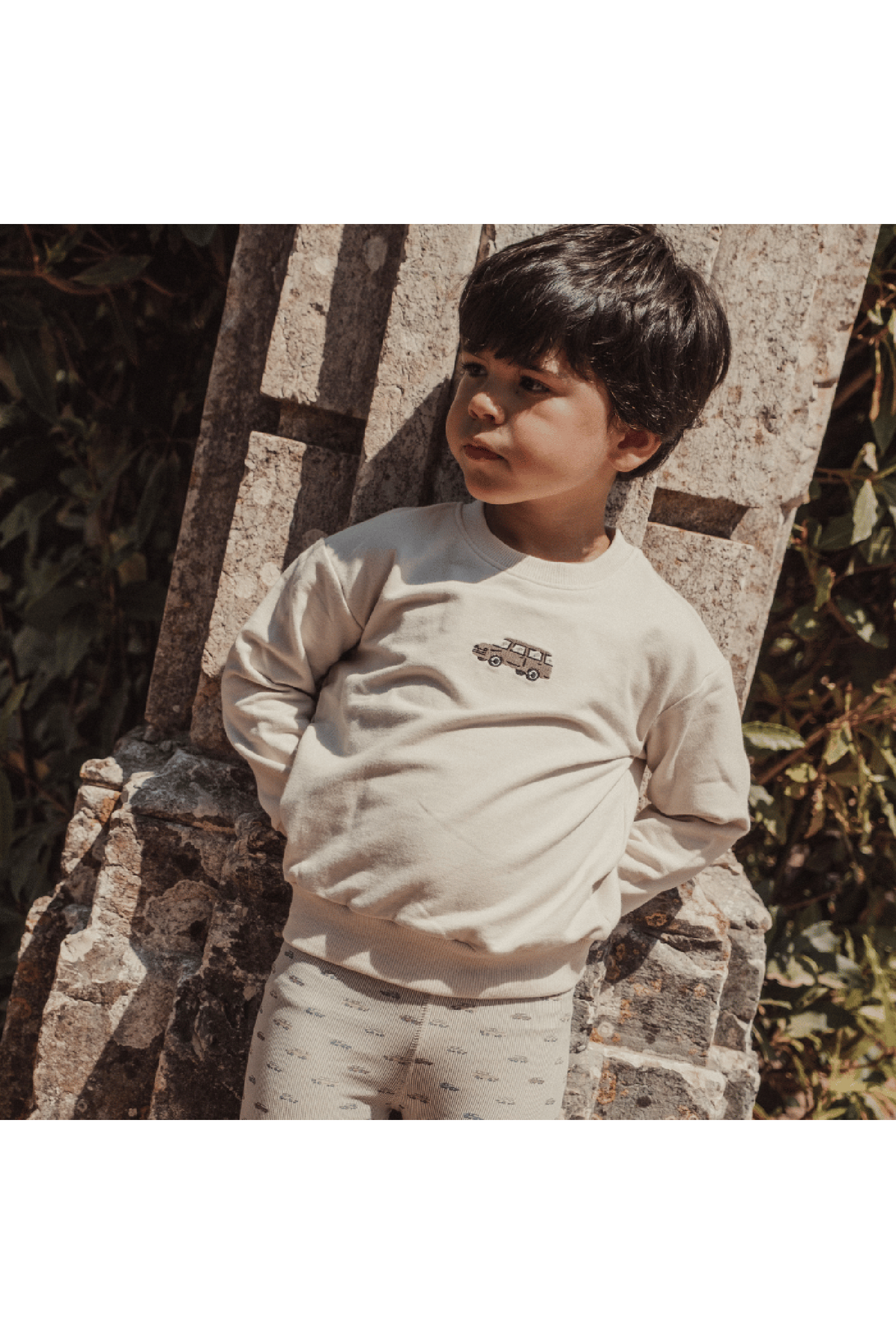 A young boy stands against a stone wall, wearing a light beige sweatshirt featuring a vintage car graphic and soft, patterned leggings.