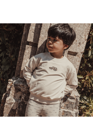 A young boy stands against a stone wall, wearing a light beige sweatshirt featuring a vintage car graphic and soft, patterned leggings.