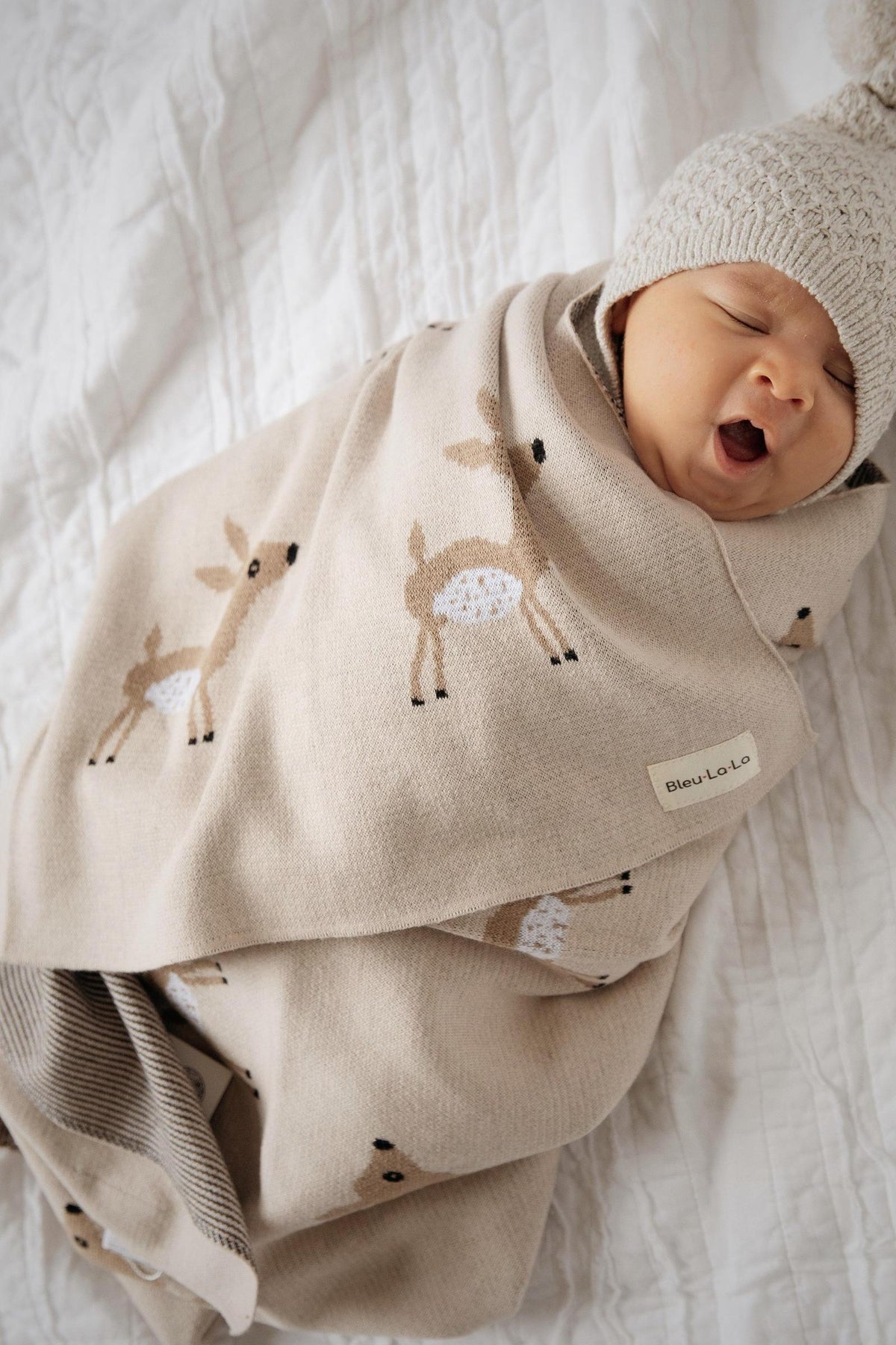 A baby wrapped in a beige swaddle blanket with deer patterns, wearing a knitted hat, lying on a white quilt.