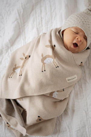 A baby wrapped in a beige swaddle blanket with deer patterns, wearing a knitted hat, lying on a white quilt.