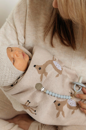 A baby wrapped in a beige knit blanket with deer design, wearing a matching knit hat and holding a pacifier.