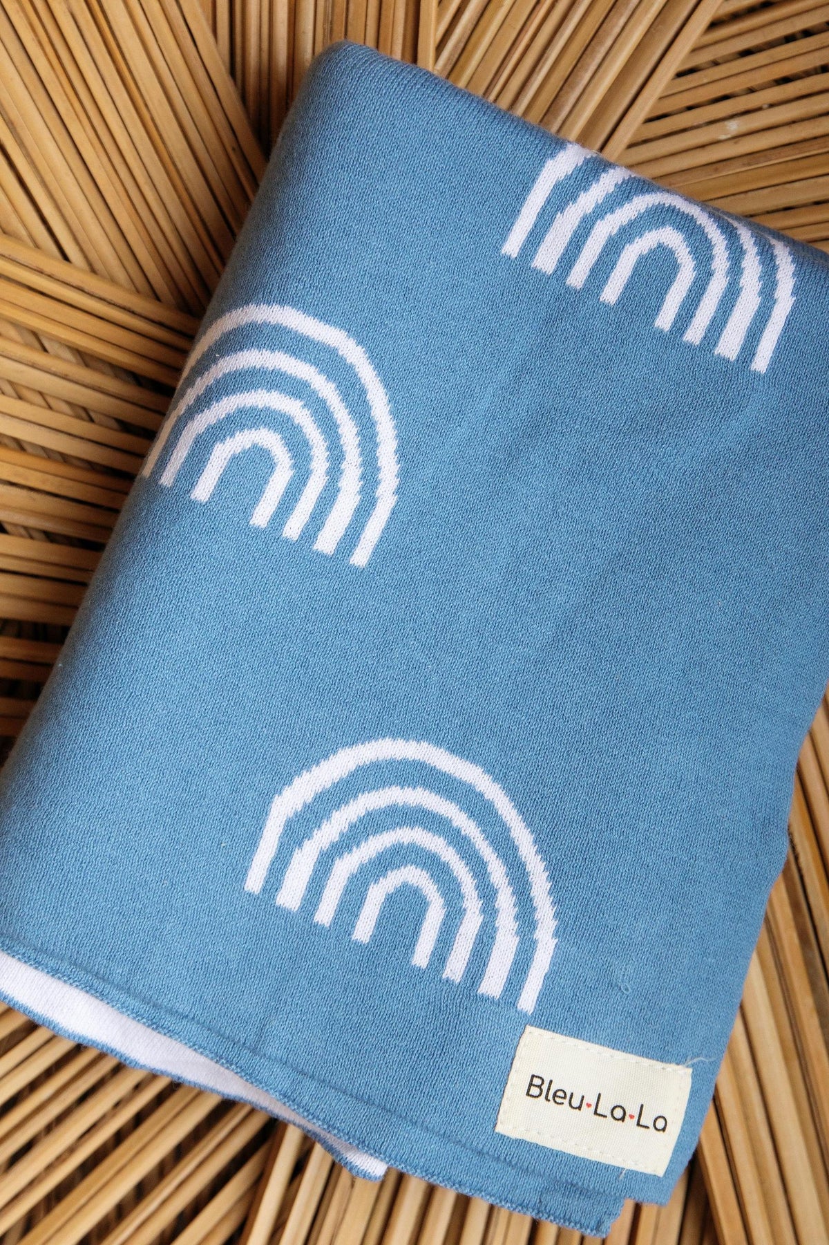 A blue swaddle blanket with white rainbow patterns, labeled Bleu-La-La, on a woven wooden surface.