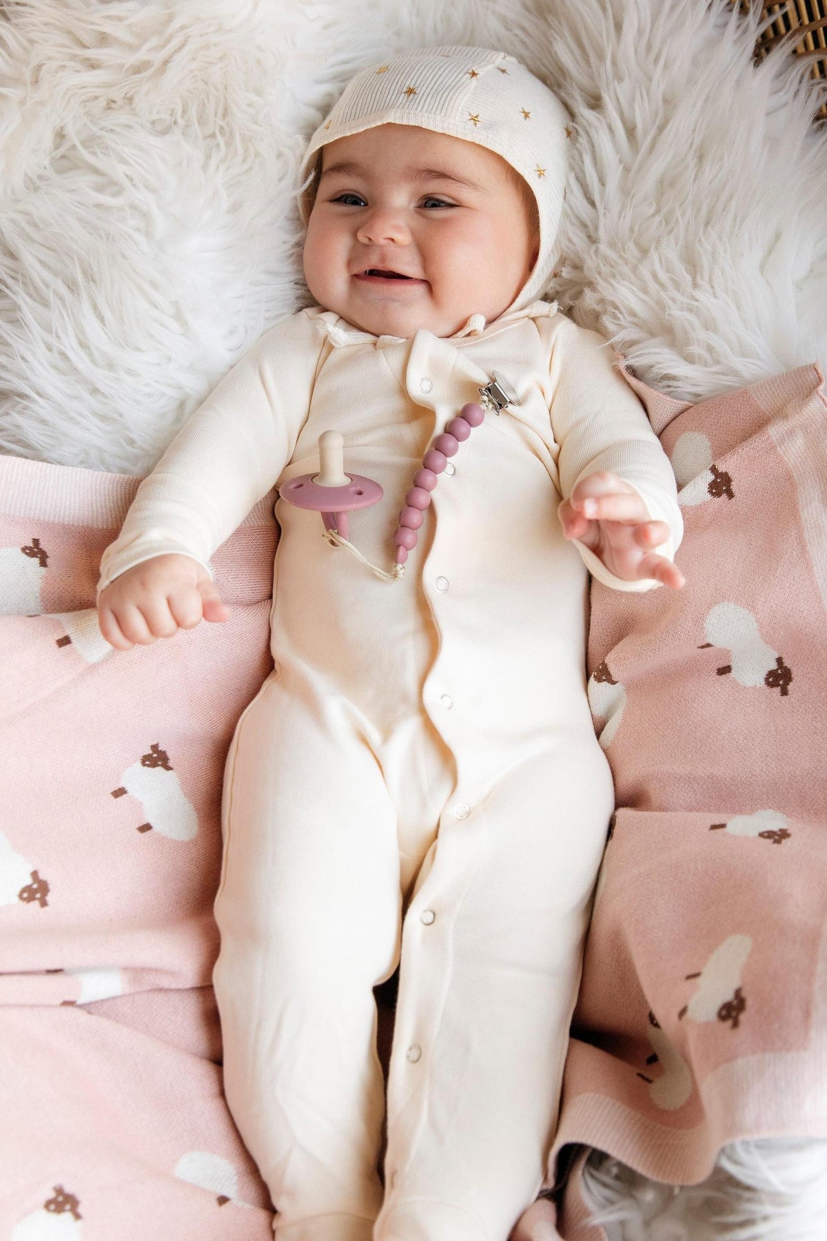 A baby in cream pajamas lies on a pink sheep-patterned blanket, smiling, with a pacifier clip attached.