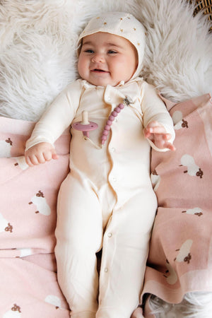 A baby in cream pajamas lies on a pink sheep-patterned blanket, smiling, with a pacifier clip attached.