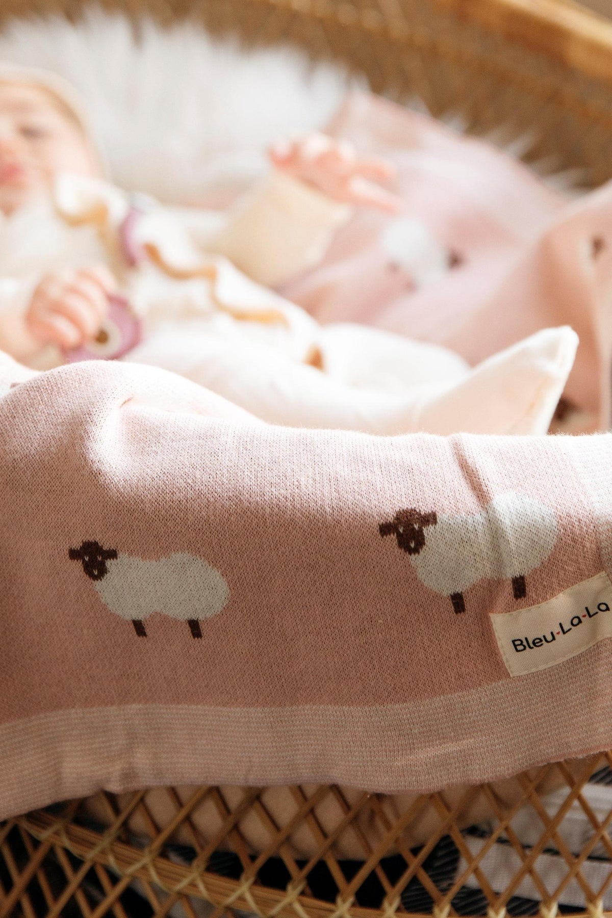 A soft pink knit blanket with sheep design draped over a baby in a wicker basket.