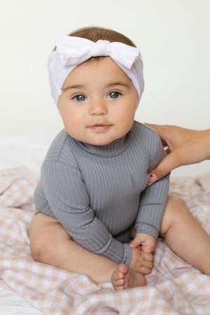 A baby wearing a gray ribbed outfit and white headband sits on a checkered blanket.