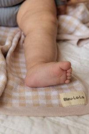 A babys leg rests on a beige and white checkered swaddle blanket with a Bleu-La-La label.