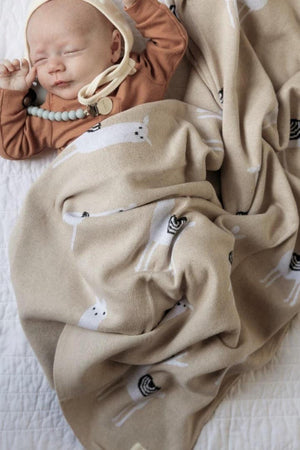 A baby sleeps under a beige llama-patterned blanket, wearing a brown outfit and cream bonnet.