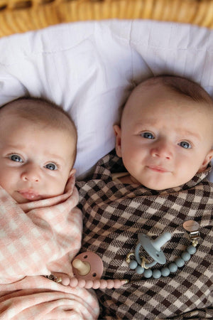 Two babies wrapped in checkered swaddle blankets, one pink and one brown, with pacifiers clipped.
