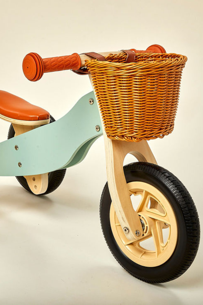 A pastel green wooden balance bike features a comfortable orange seat and a woven basket.