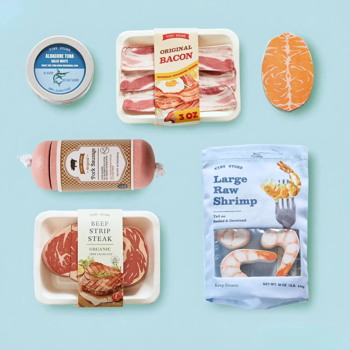 A colorful assortment of play food items includes wooden representations of bacon, tuna, shrimp, and steak, all presented on a light blue background.