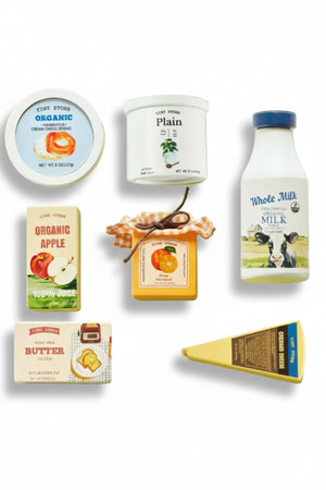An assortment of wooden play food items on a light green background, featuring organic milk, juice, butter, and cheese, all with colorful, playful packaging designed for imaginative play.