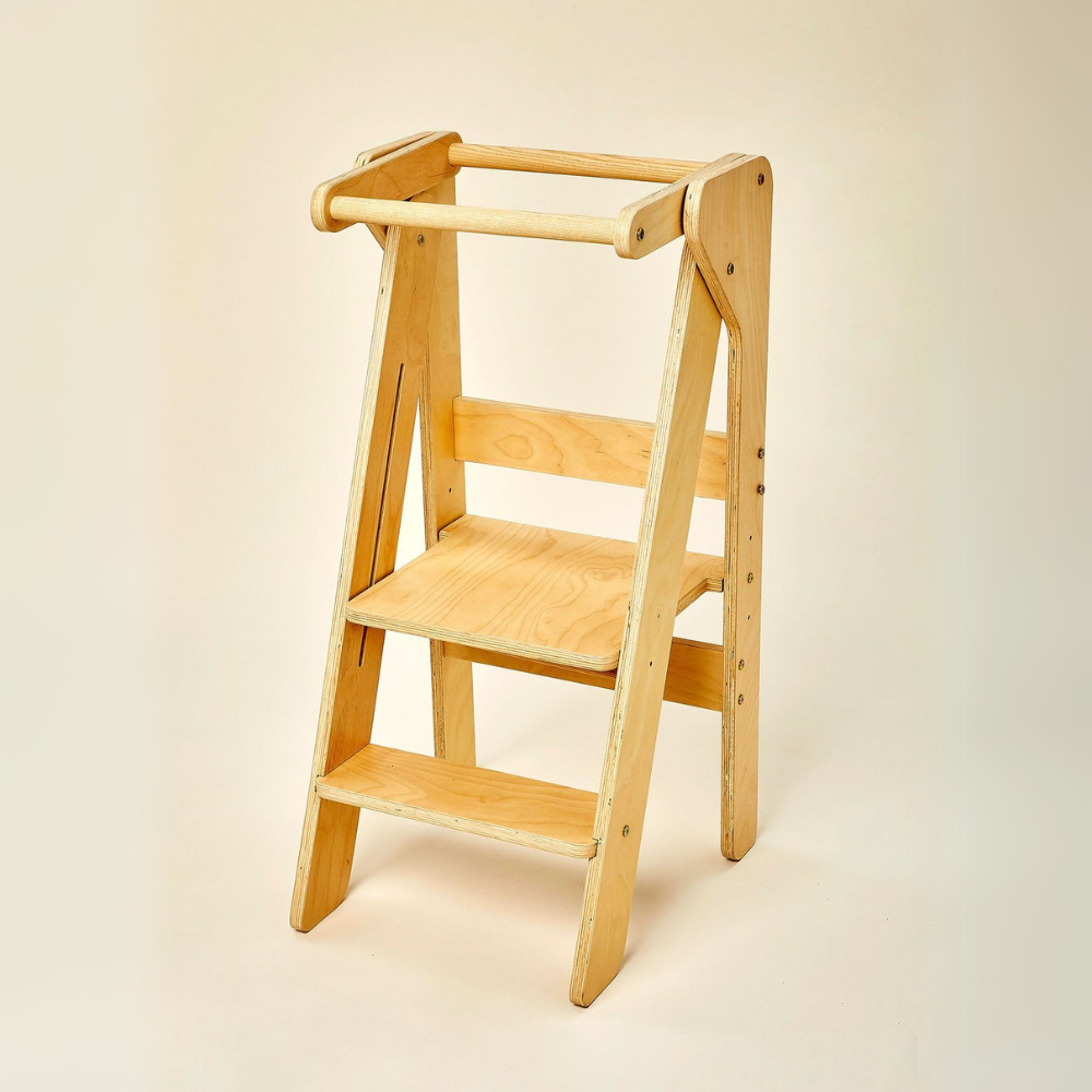 A natural wood foldable educational tower features a sturdy design with two steps and a safety rail, ideal for encouraging children to participate in cooking and other activities alongside adults.