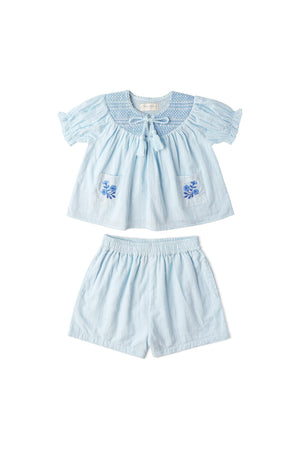 Light blue organic cotton smocked top and shorts set featuring delicate embroidery on the pockets. This playful outfit is perfect for sunny day outings for young children.