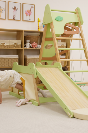 A child plays underneath a green wooden climber featuring a slide, ladder, and tunnel. The indoor setting includes storage baskets and framed animal illustrations on the wall.