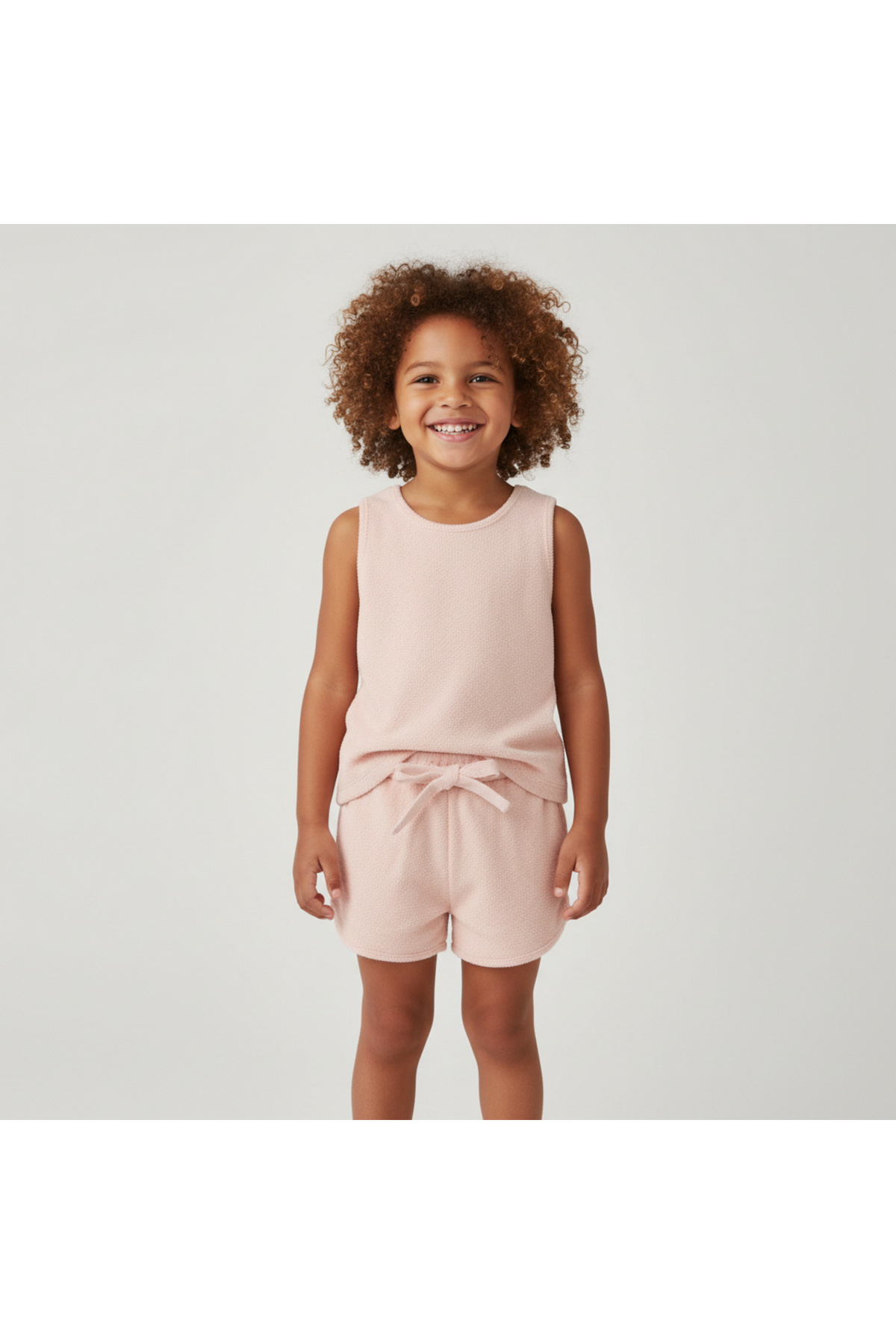A young child with curly hair smiles while wearing a blush-colored organic sleeveless tee and matching shorts set, made from soft terry material, perfect for comfortable play.