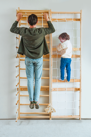 A man performs a pull-up on a wooden Swedish wall ladder while a child plays on an adjacent climbing structure.