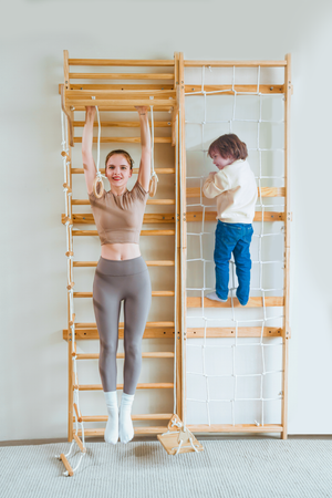 A woman performs an exercise on a wooden Swedish wall ladder while a child climbs a rope net.