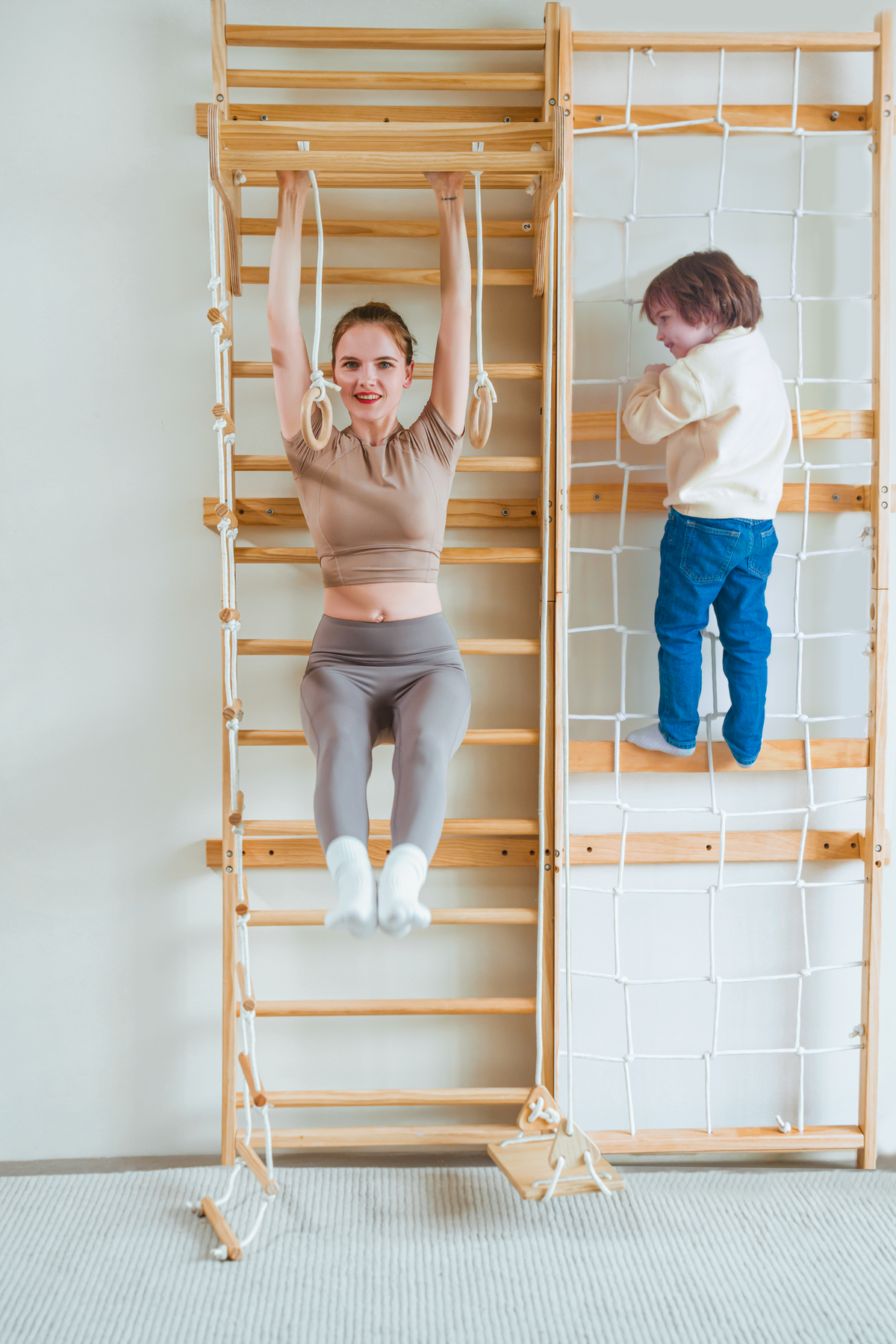 A woman performs a hanging exercise on a wooden Swedish wall ladder, while a child climbs a rope wall nearby.