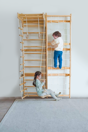 A wooden Swedish wall ladder features a child climbing and another sitting on a swing, both engaged in play indoors.