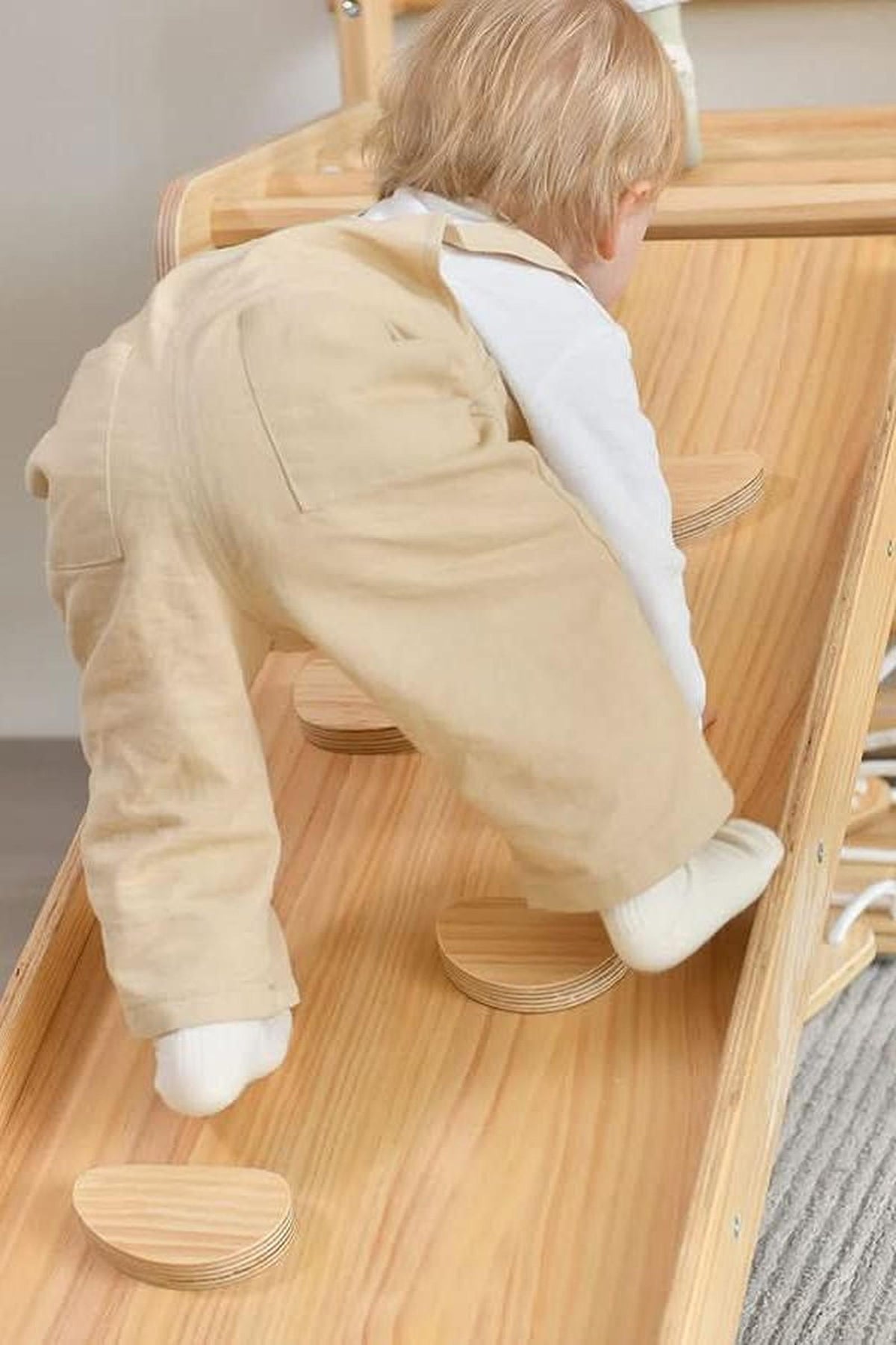 A toddler in beige overalls climbs a wooden play structure with circular steps, showcasing natural wood grain and a safe.