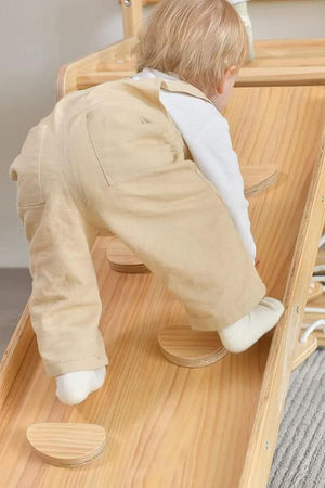 A toddler in beige overalls climbs a wooden play structure with circular steps, showcasing natural wood grain and a safe.