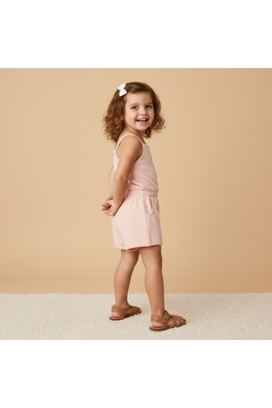 A young girl smiles while wearing a soft blush tank romper with a cinched waist, paired with brown sandals. The backdrop features a warm, neutral tone, enhancing the playful vibe.