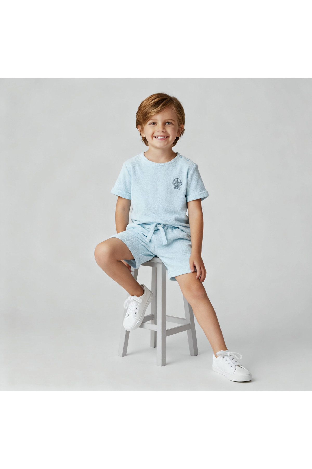 A young child smiles while sitting on a gray stool, wearing a light blue organic terry tee and shorts set. The outfit features a relaxed fit and soft, breathable fabric.