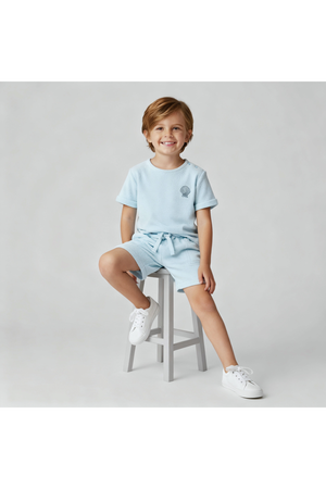 A young child smiles while sitting on a gray stool, wearing a light blue organic terry tee and shorts set. The outfit features a relaxed fit and soft, breathable fabric.