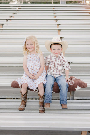 A boy and girl sit on a bleacher, dressed in western attire. The boy wears a patterned button-up shirt and cowboy hat, while the girl is in a floral dress.