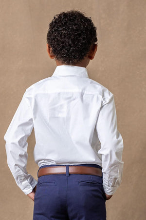A child wearing a white organic cotton button-down shirt stands with their back to the camera. The shirt features a classic collar and is paired with navy blue pants.