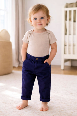 A toddler stands barefoot, wearing a light beige ribbed onesie and navy chinos with rolled cuffs, smiling in a cozy.