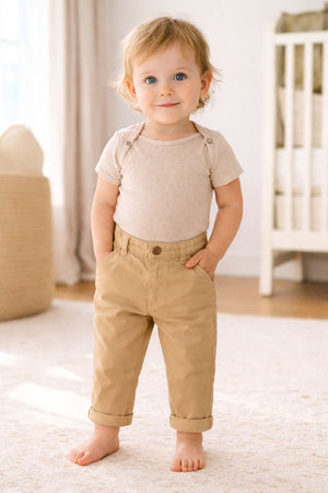 A toddler stands smiling, wearing a beige ribbed onesie and khaki chinos, with hands in pockets, on a light rug.