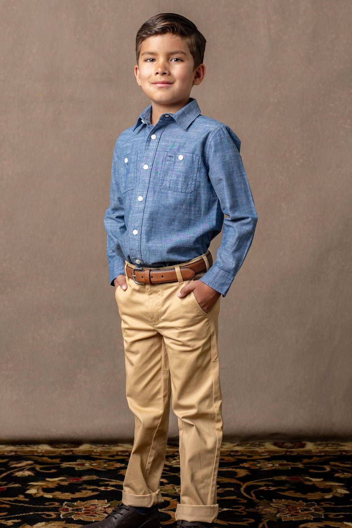 A boy stands confidently in a blue button-up shirt and khaki chinos, with hands in his pockets, on a patterned.
