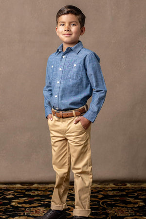 A boy stands confidently in a blue button-up shirt and khaki chinos, with hands in his pockets, on a patterned.