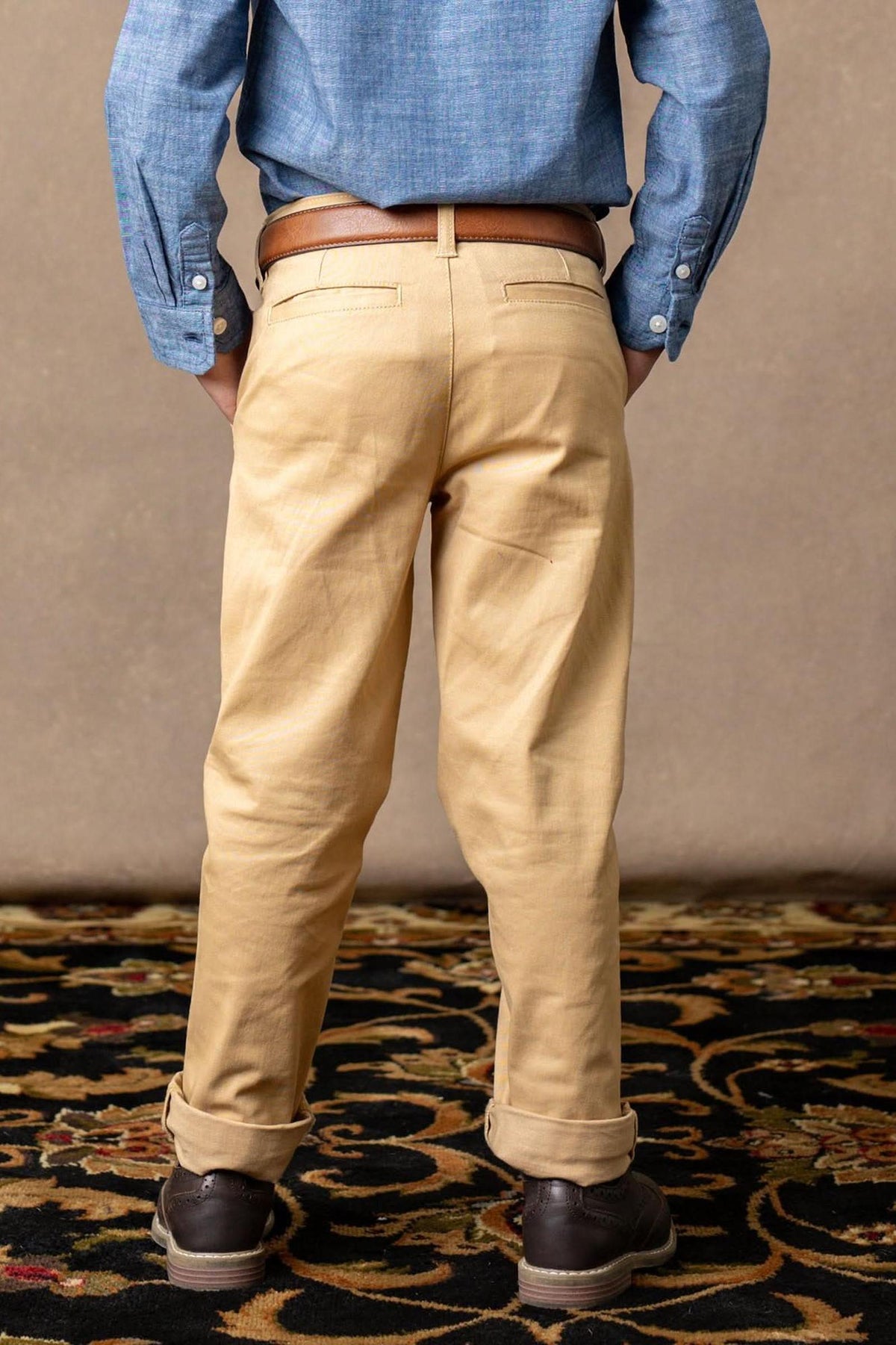 A boy is wearing khaki skinny chinos, paired with a blue shirt and brown shoes, standing on an ornate rug.